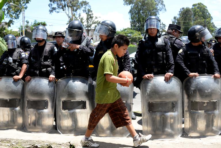 Một đứa trẻ ôm quả bóng đi ngang qua hàng hàng rào cảnh sát chống bạo động Guatemala, nơi nhà chức trách đã cho khoảng 200 gia đình lấn chiếm đất 15 ngày để sơ tán khỏi nơi công cộng mà họ đã chiếm giữ 2 năm qua