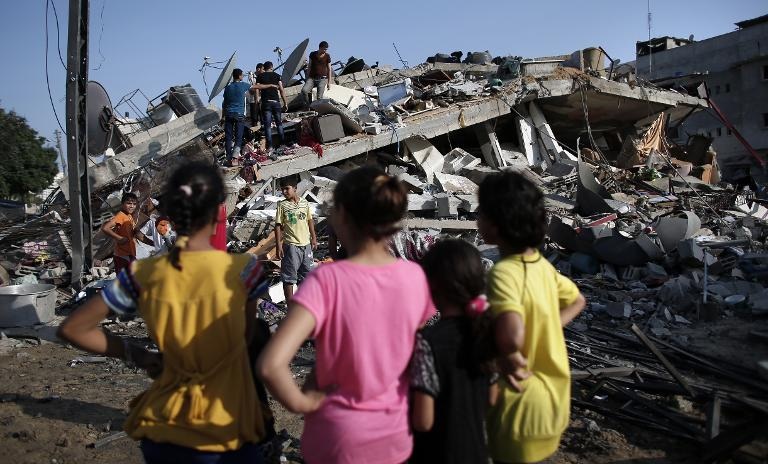 Những người Palestine đứng trước một tòa nhà đã bị phá hủy bởi một cuộc không kích của Israel tại thành phố Gaza
