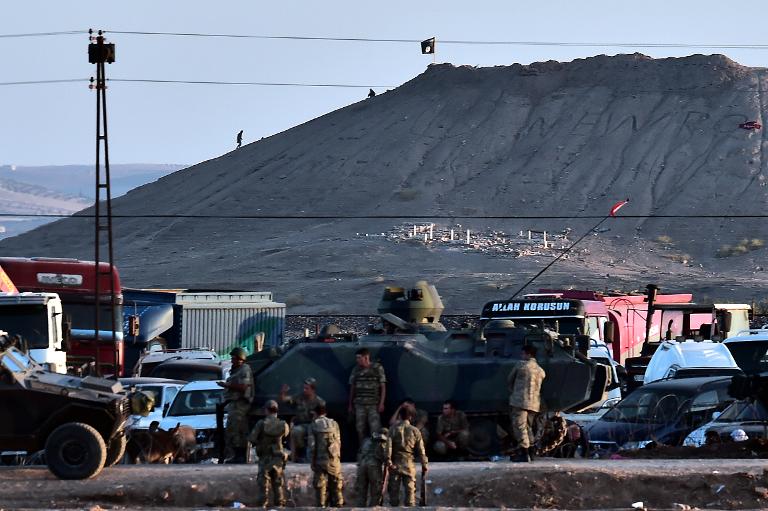Một nhóm chiến binh đứng phía dưới ngọn đồi có cắm cờ của tổ chức IS, phía ngoài thị trấn Kobane
