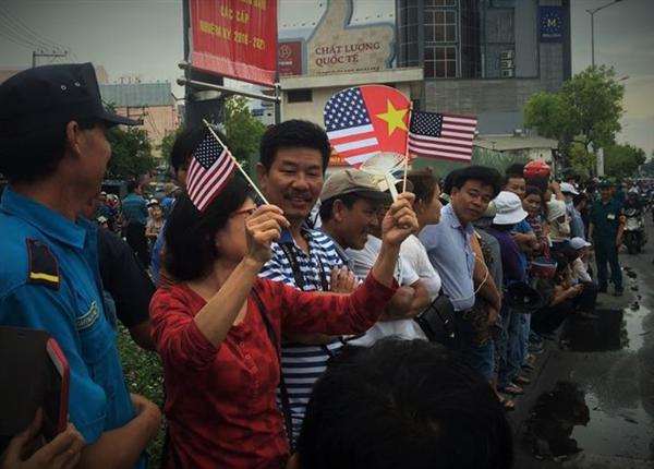 Một người dân cầm cờ Việt Nam và cờ Hoa Kỳ đứng chờ đoàn xe đi qua.