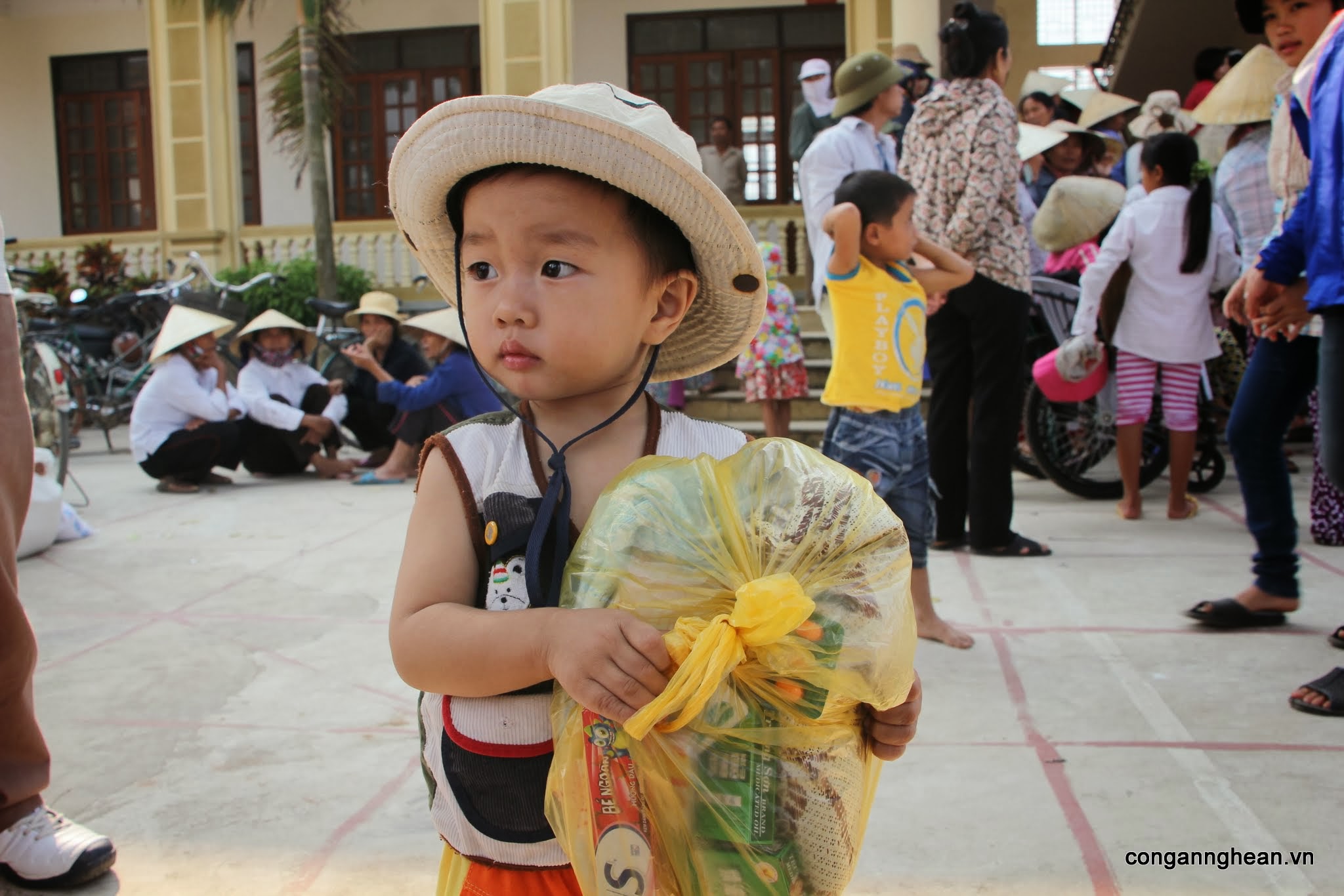 Bé Hoàng Đức Thiện, 2 tuổi, tay ôm gói quà, ngơ ngác trước quá đông người, trong lúc mẹ vào nhận gạo.