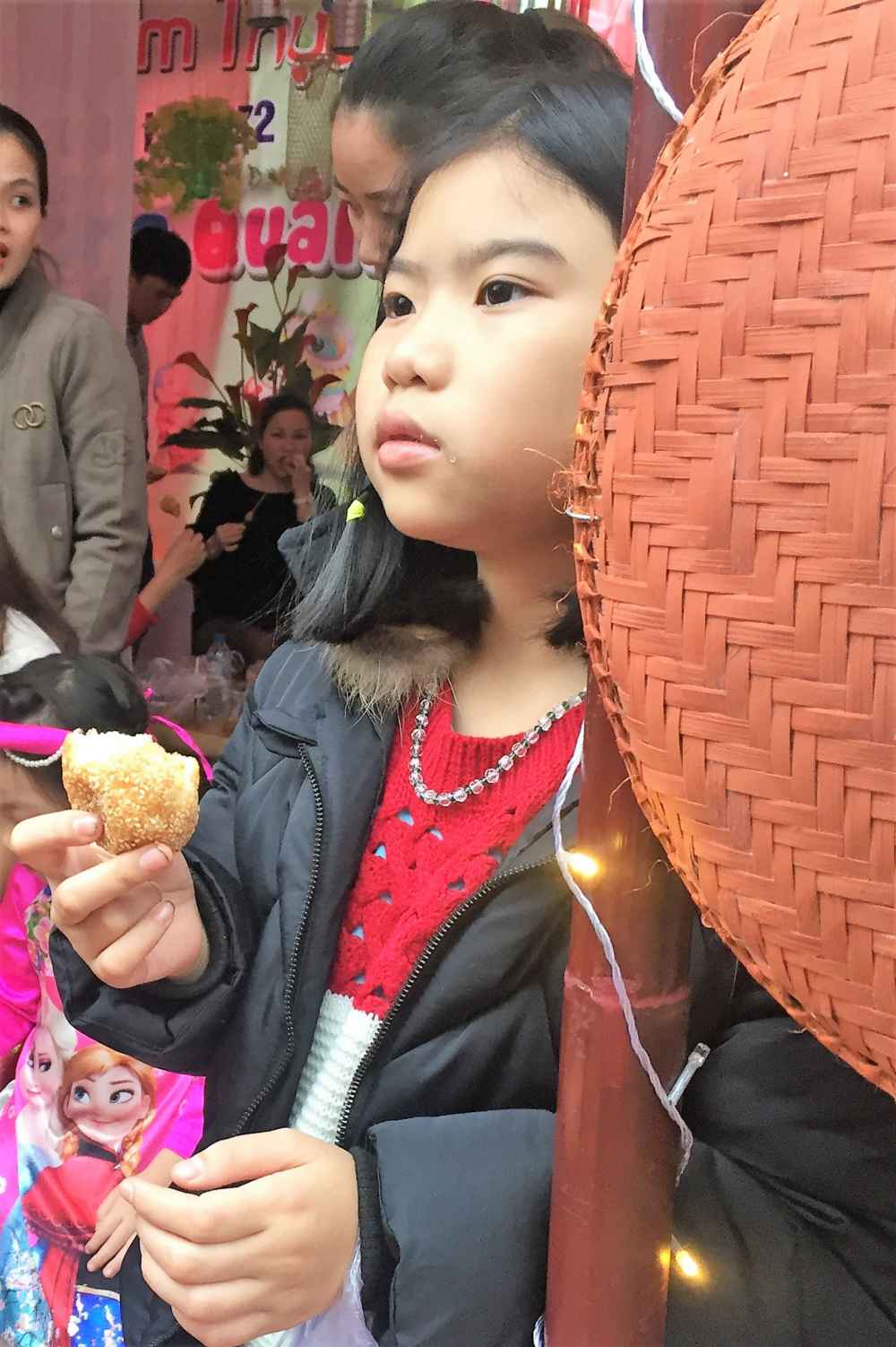 Một cô bé ngắm dòng người qua lại, trên tay vẫn cầm chiếc bánh rán quê